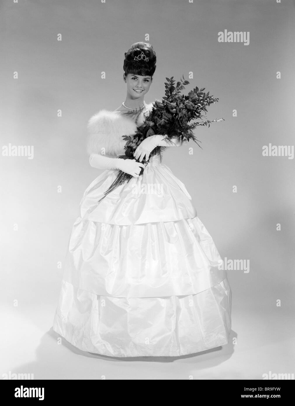 1960s PRETTY YOUNG WOMAN IN EVENING DRESS AT  BEAUTY PAGEANT WEARING FUR STOLE AND TIARA HOLDING BOUQUET OF ROSES LOOKING AT Stock Photo