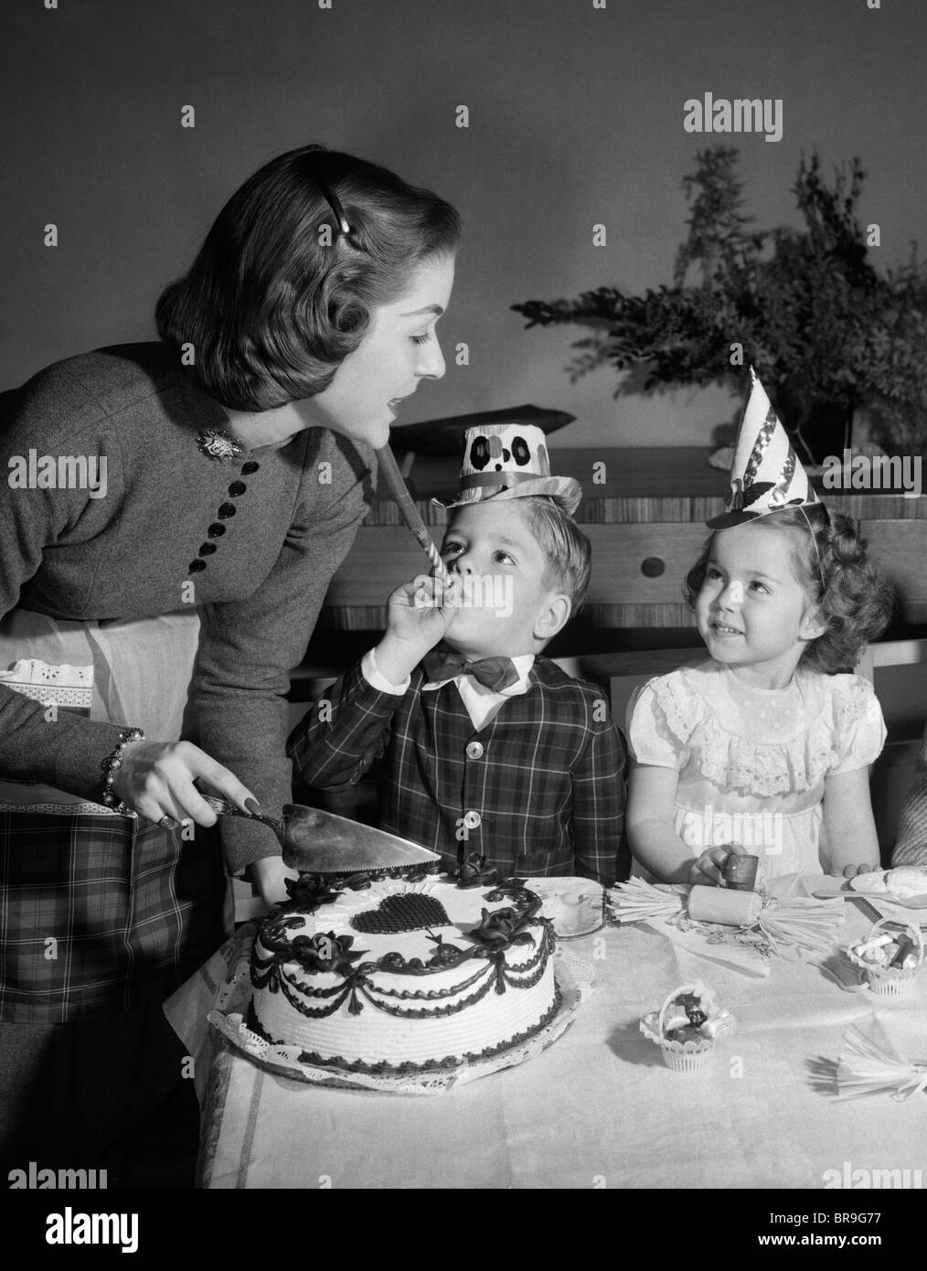 1950s WOMAN MOTHER CUTTING BIRTHDAY CAKE FOR TWO CHILDREN SITTING AT TABLE WEARING PARTY HATS Stock Photo