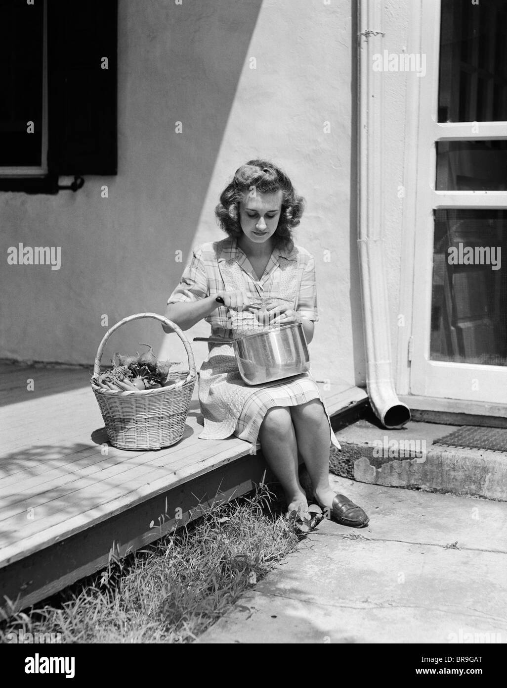 1940s WOMAN WEARING PLAID DRESS AND APRON SITTING ON PORCH OF FARMHOUSE WITH BASKET OF VEGETABLES PEELING TOMATO OVER POT Stock Photo