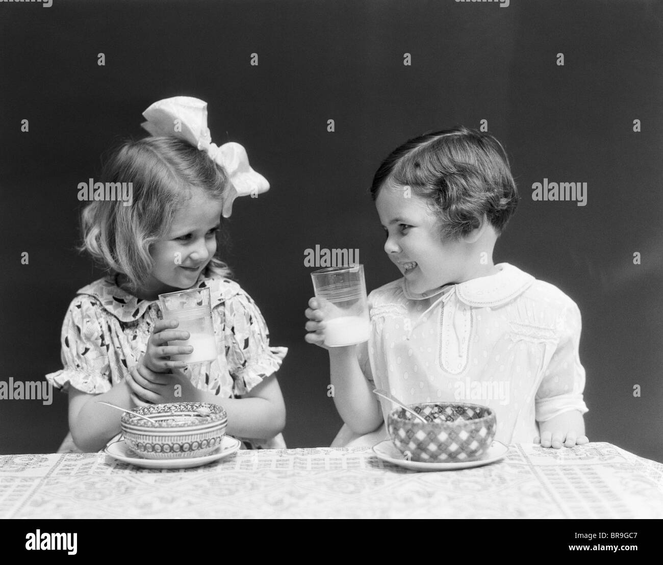 1930s TWO SMILING LITTLE GIRLS DRINKING MILK BOWL OF CEREAL TALKING LAUGHING EATING Stock Photo