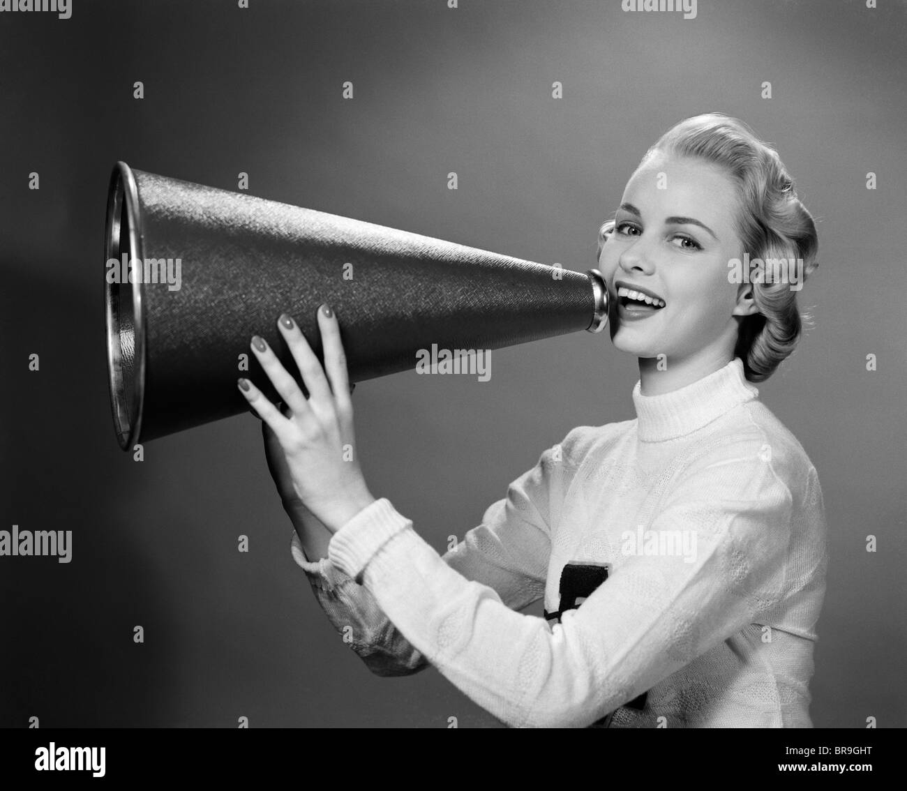 1950s BLONDE WOMAN CHEERLEADER YELLING INTO MEGAPHONE LOOKING AT CAMERA Stock Photo