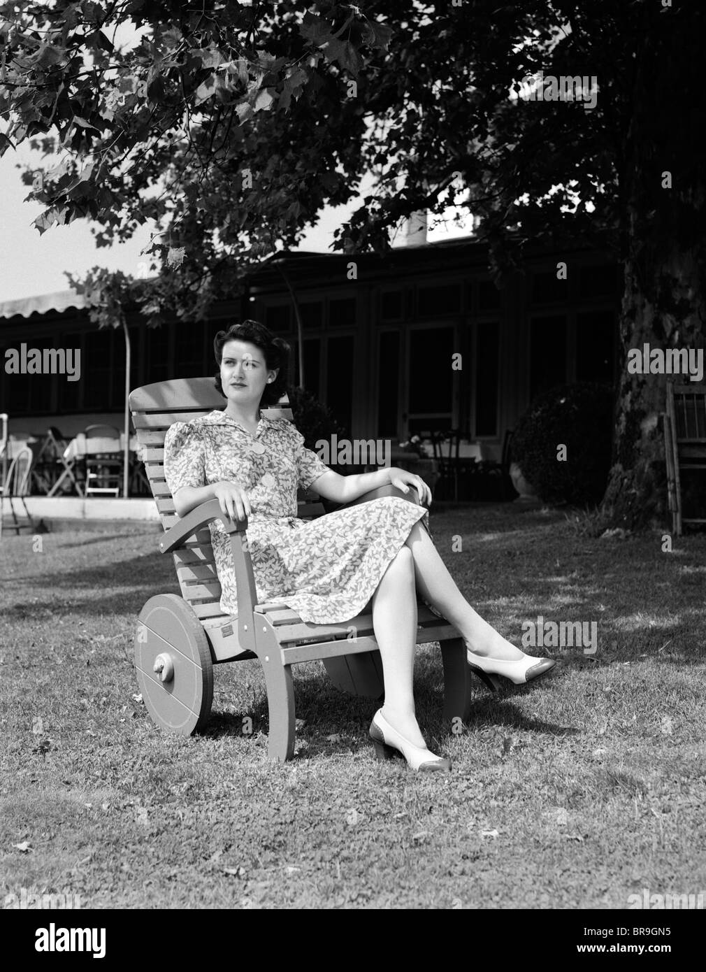 1940s WOMAN SITTING IN WOODEN LAWN CHAIR WITH WHEELS ON LAWN IN FRONT OF HOUSE LOOKING OFF INTO DISTANCE Stock Photo