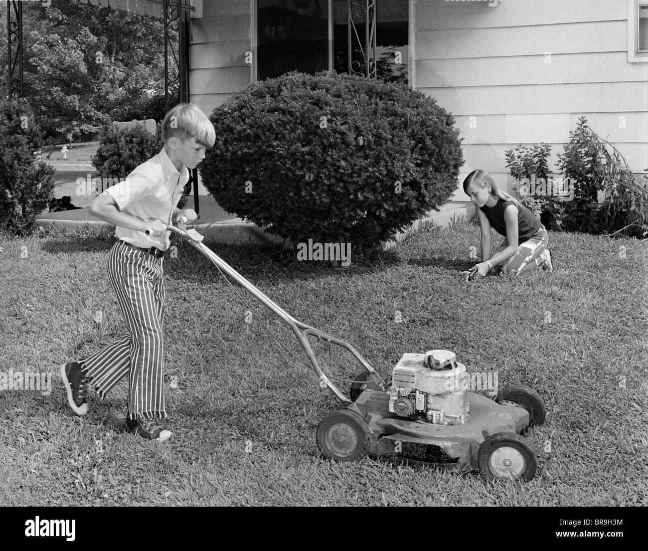 1970s BROTHER AND SISTER DOING CHORES MOWING LAWN CUTTING GRASS YARD WORK TOGETHER Stock Photo