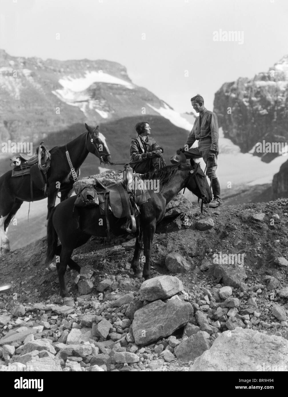 1920s 1930s COUPLE MAN WOMAN WEARING RIDING GEAR JODHPURS BOOTS SPURS SITTING STANDING ON LARGE ROCK BY TWO HORSES Stock Photo
