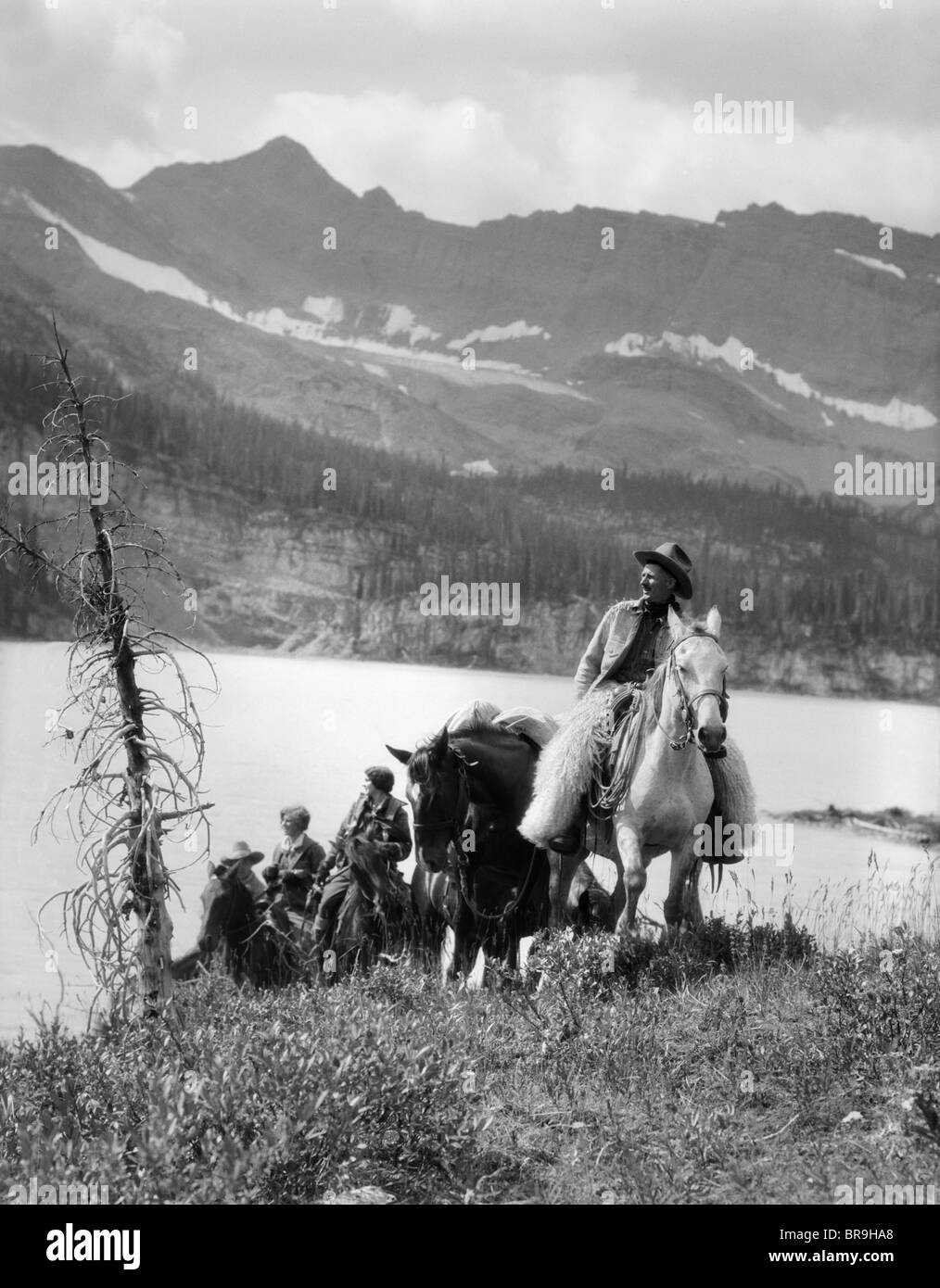 1920s 1930s TWO WOMEN ONE MAN RIDING HORSES WESTERN SADDLES COWBOY WEARING WOOL CHAPS BY MOUNTAIN LAKE IN CANADA Stock Photo