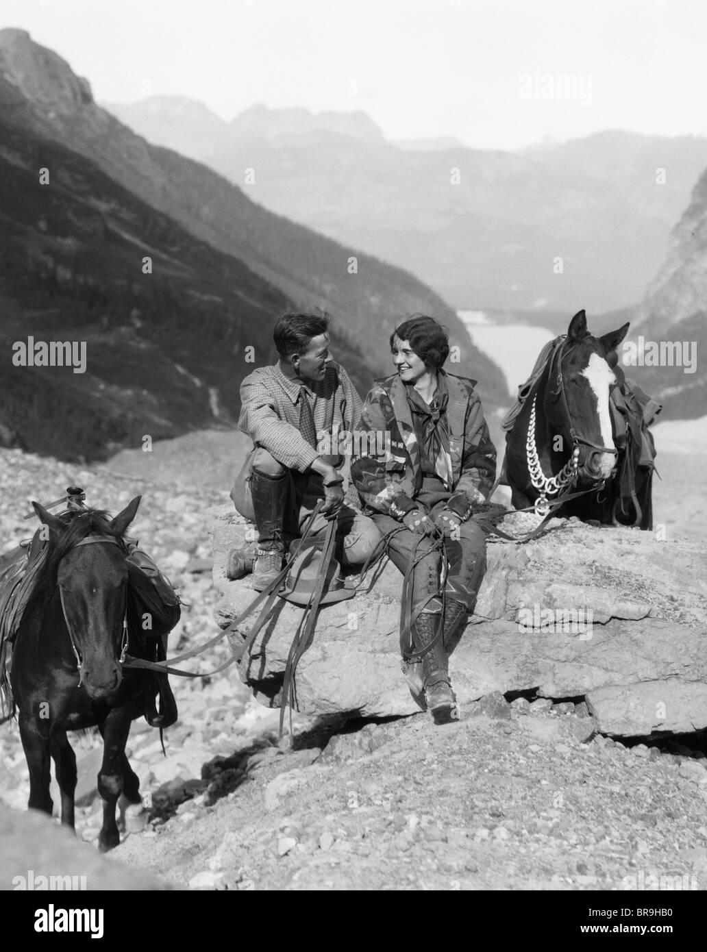 1920s 1930s COUPLE MAN WOMAN WEARING RIDING GEAR JODHPURS BOOTS SPURS SITTING ON LARGE ROCK BY TWO HORSES WITH WESTERN SADDLES Stock Photo