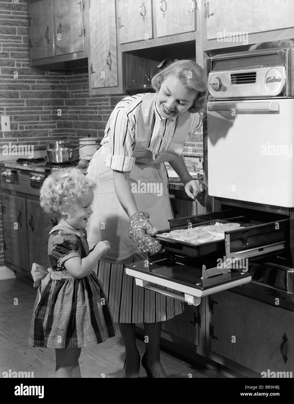 1950s SMILING MOTHER DAUGHTER IN KITCHEN BROILING PORK CHOPS Stock Photo