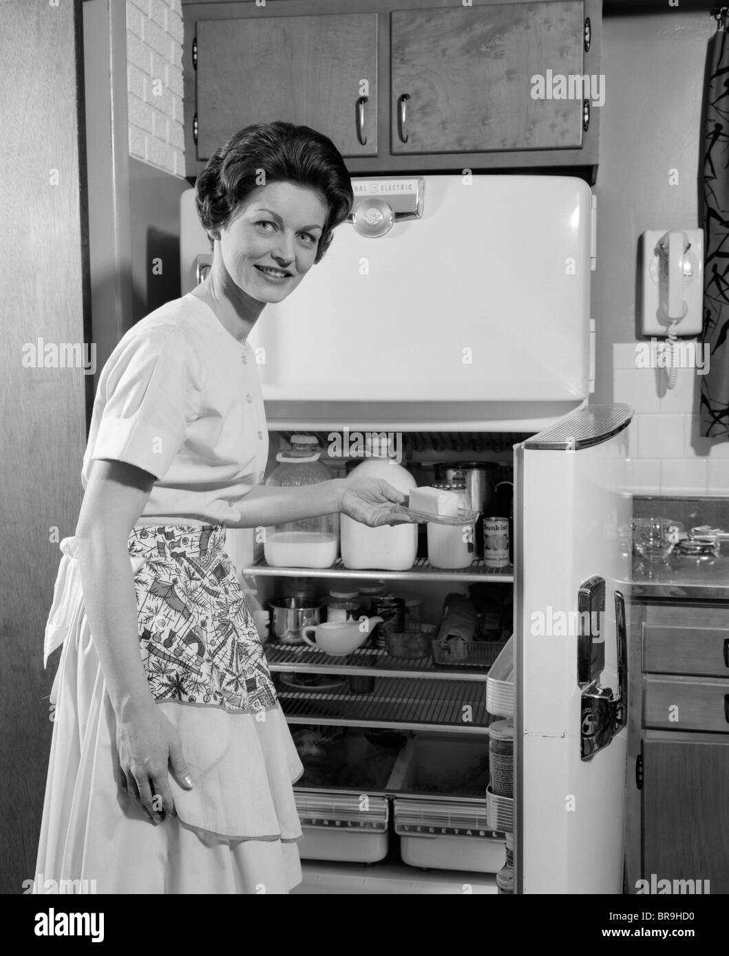 1950s SMILING WOMAN HOUSEWIFE PUTTING STICK OF BUTTER INTO ELECTRIC REFRIGERATOR IN KITCHEN LOOKING AT CAMERA Stock Photo