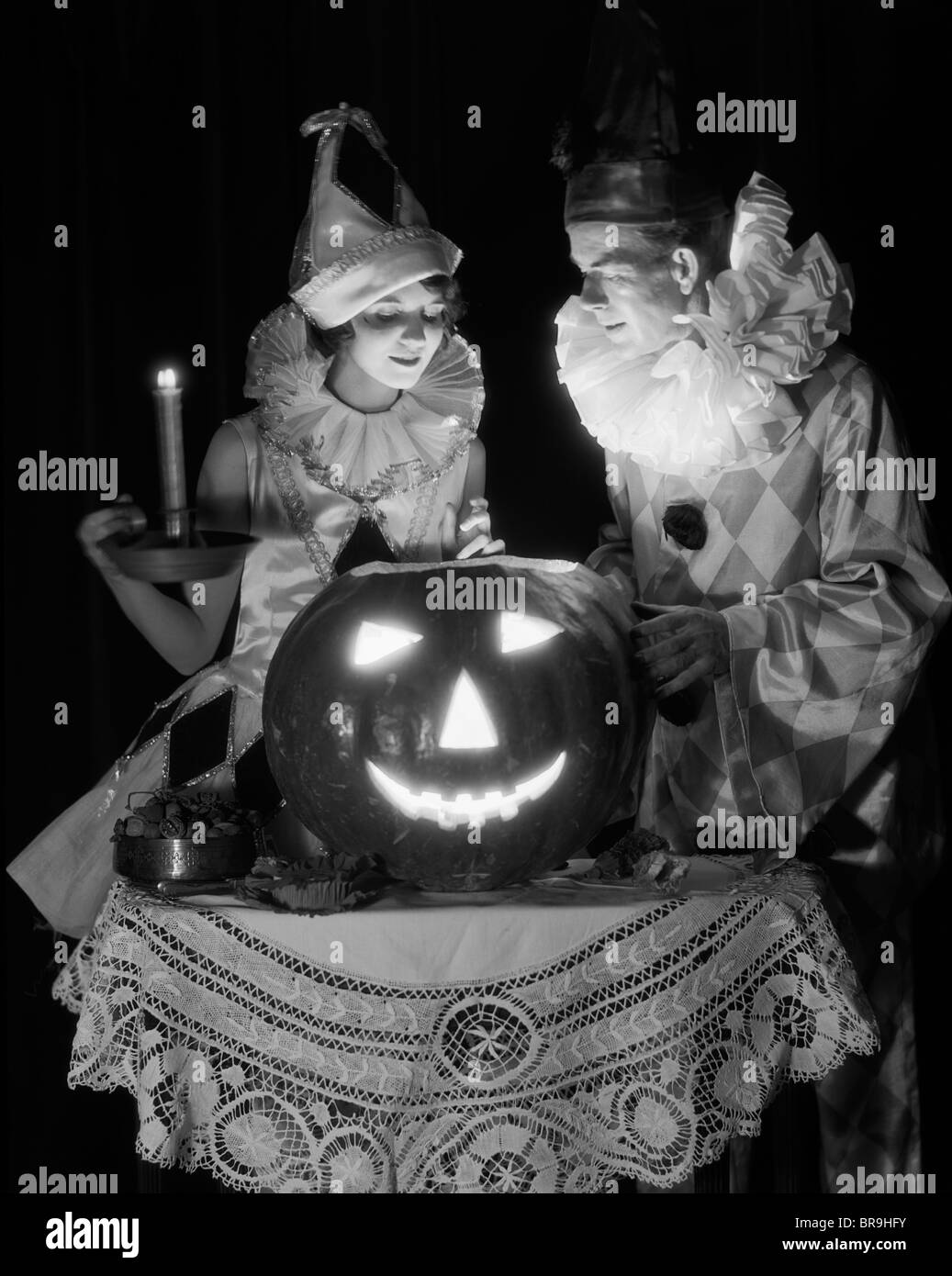 1920s 1930s COUPLE IN COSTUMES LOOKING DOWN INTO CANDLE LIGHT FROM CARVED PUMPKIN JACK-O-LANTERN Stock Photo
