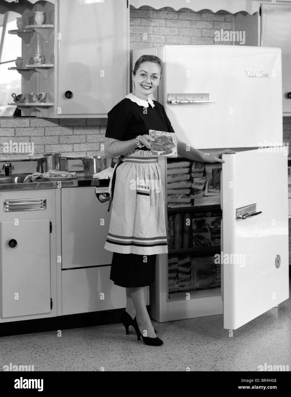 1950s SMILING WOMAN HOUSEWIFE IN KITCHEN TAKING FROZEN FOOD OUT OF REFRIGERATOR FREEZER Stock Photo