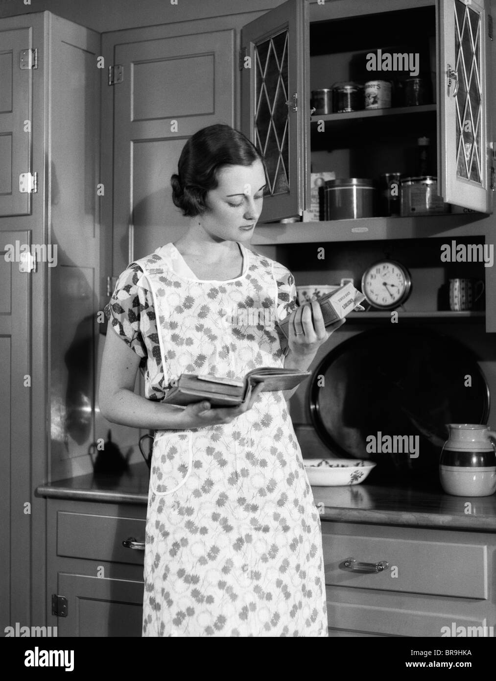 1920s 1930s WOMAN HOUSEWIFE WEARING APRON IN KITCHEN HOLDING COOKBOOK READING PACKAGE OF CORN STARCH Stock Photo