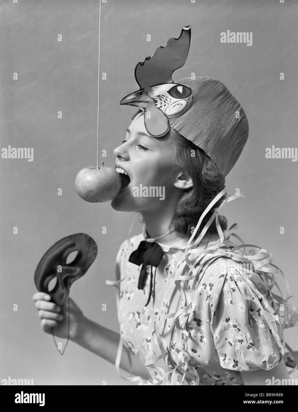 1930s 1940s GIRL BOBBING FOR APPLE DANGLING ON A STRING WEARING PARTY HAT AND HOLDING EYE MASK Stock Photo