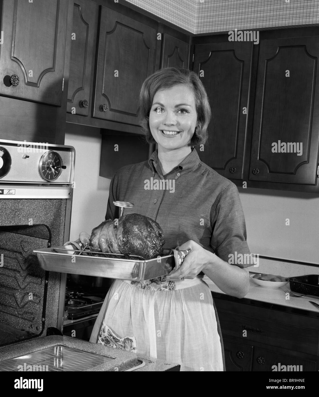 1960s SMILING WOMAN IN KITCHEN TAKING ROAST OUT OF OVEN LOOKING AT CAMERA Stock Photo