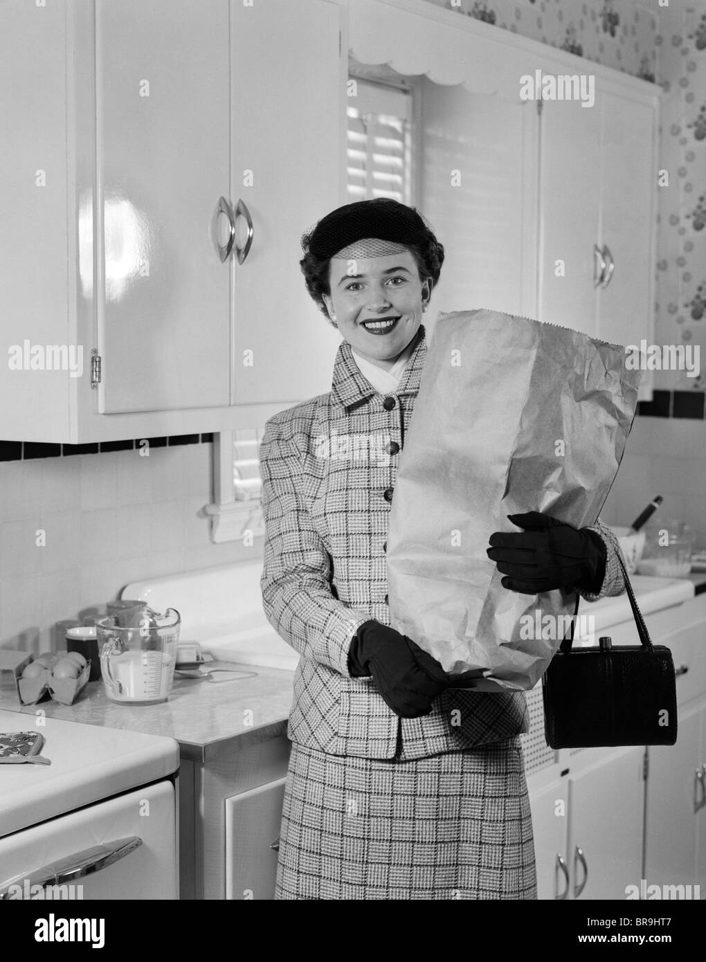 1950s SMILING WOMAN IN KITCHEN HOLDING GROCERY BAG HANDBAG WEARING HAT GLOVES LOOKING AT CAMERA Stock Photo