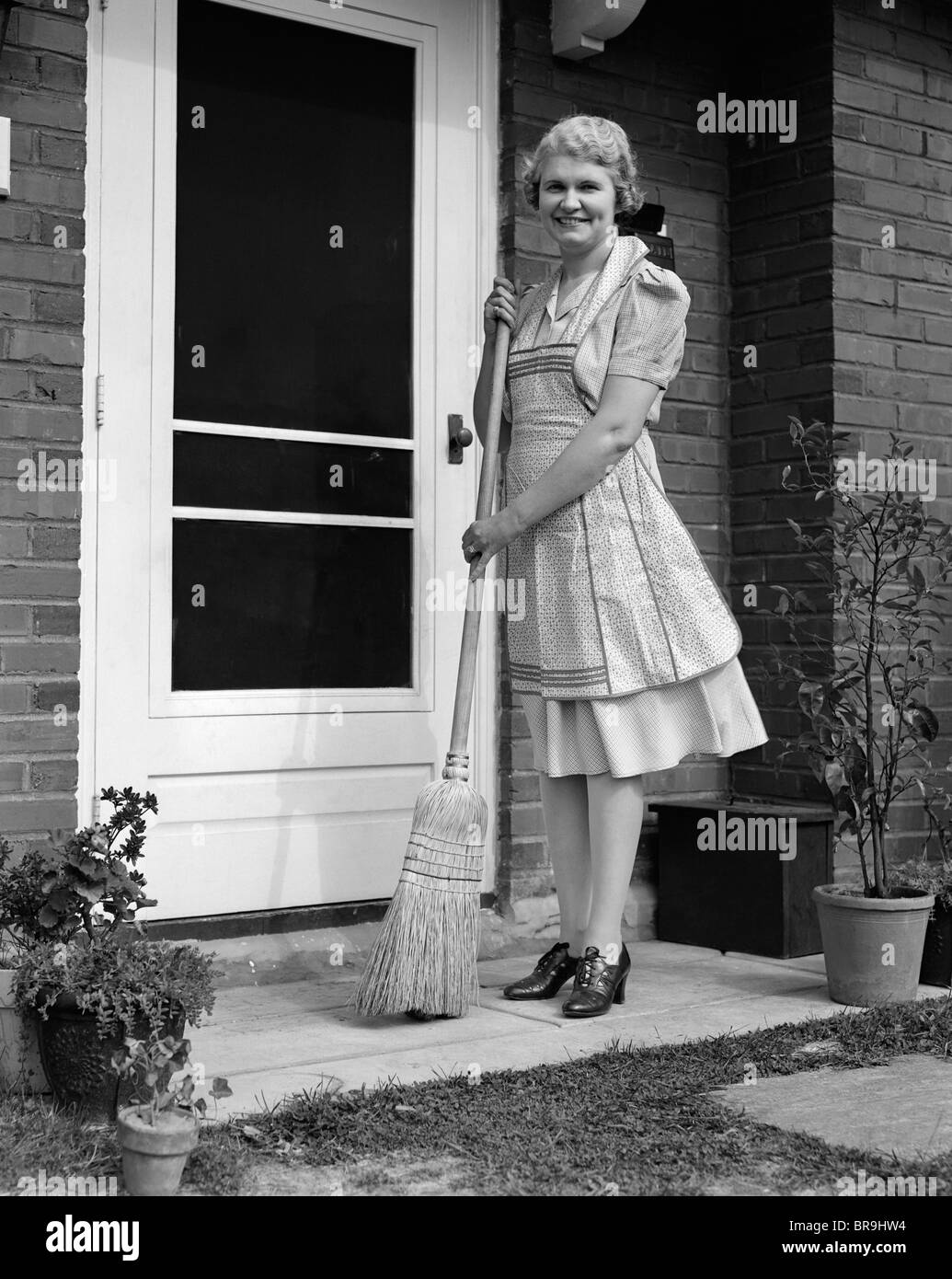 1940s SMILING WOMAN SWEEPING PORCH FRONT DOOR STEP WITH A BROOM LOOKING
