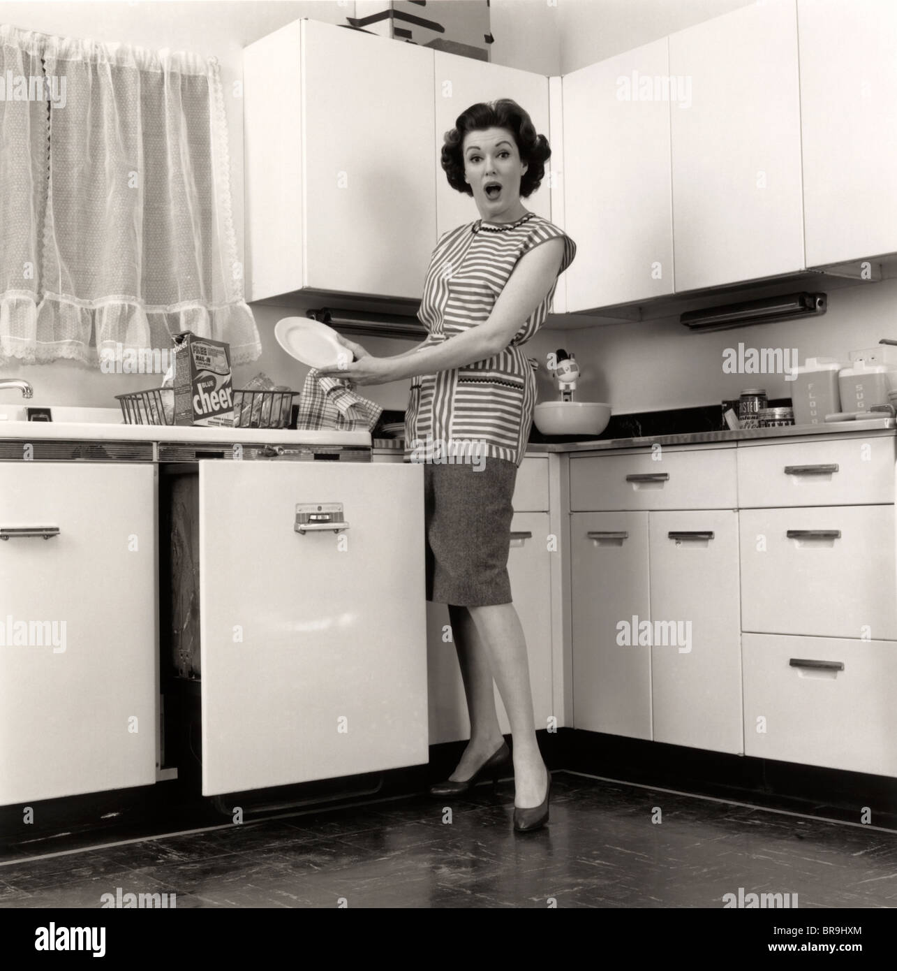 1950s WOMAN HOUSEWIFE WEARING SMOCK PUTTING DISHES IN AUTOMATIC DISHWASHER IN SUBURBAN KITCHEN LOOKING AT CAMERA Stock Photo