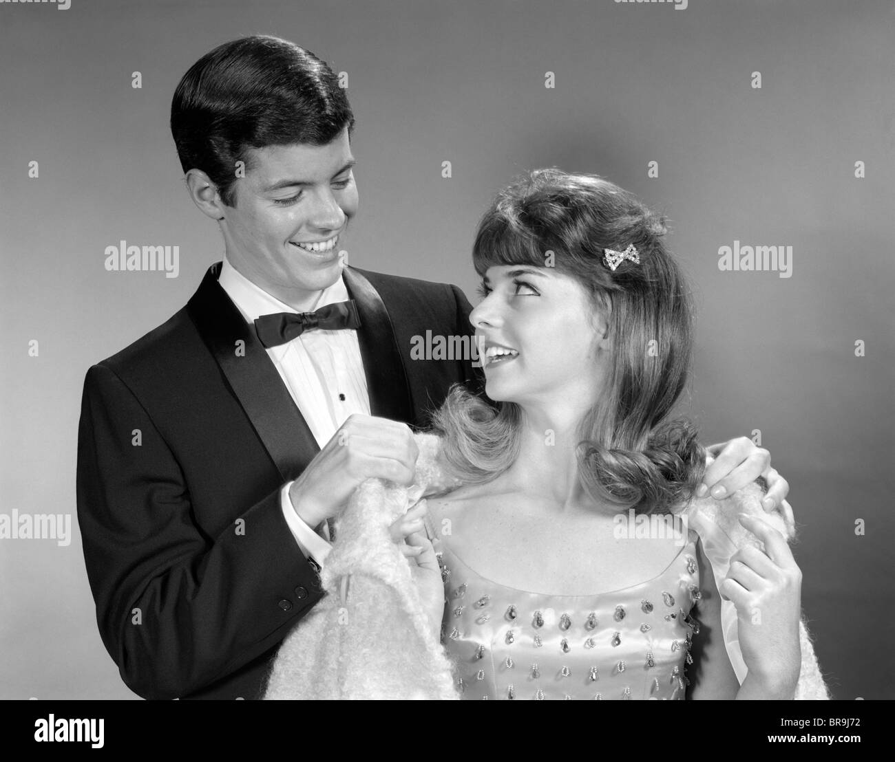 1960s TEEN COUPLE FORMALLY DRESSED BOY HELPING GIRL ON WITH COAT TO GO TO PROM Stock Photo