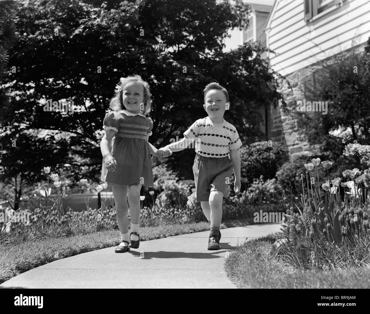 1950s TWO CHILDREN HOLDING HANDS SMILING WALKING ALONG SIDEWALK Stock Photo