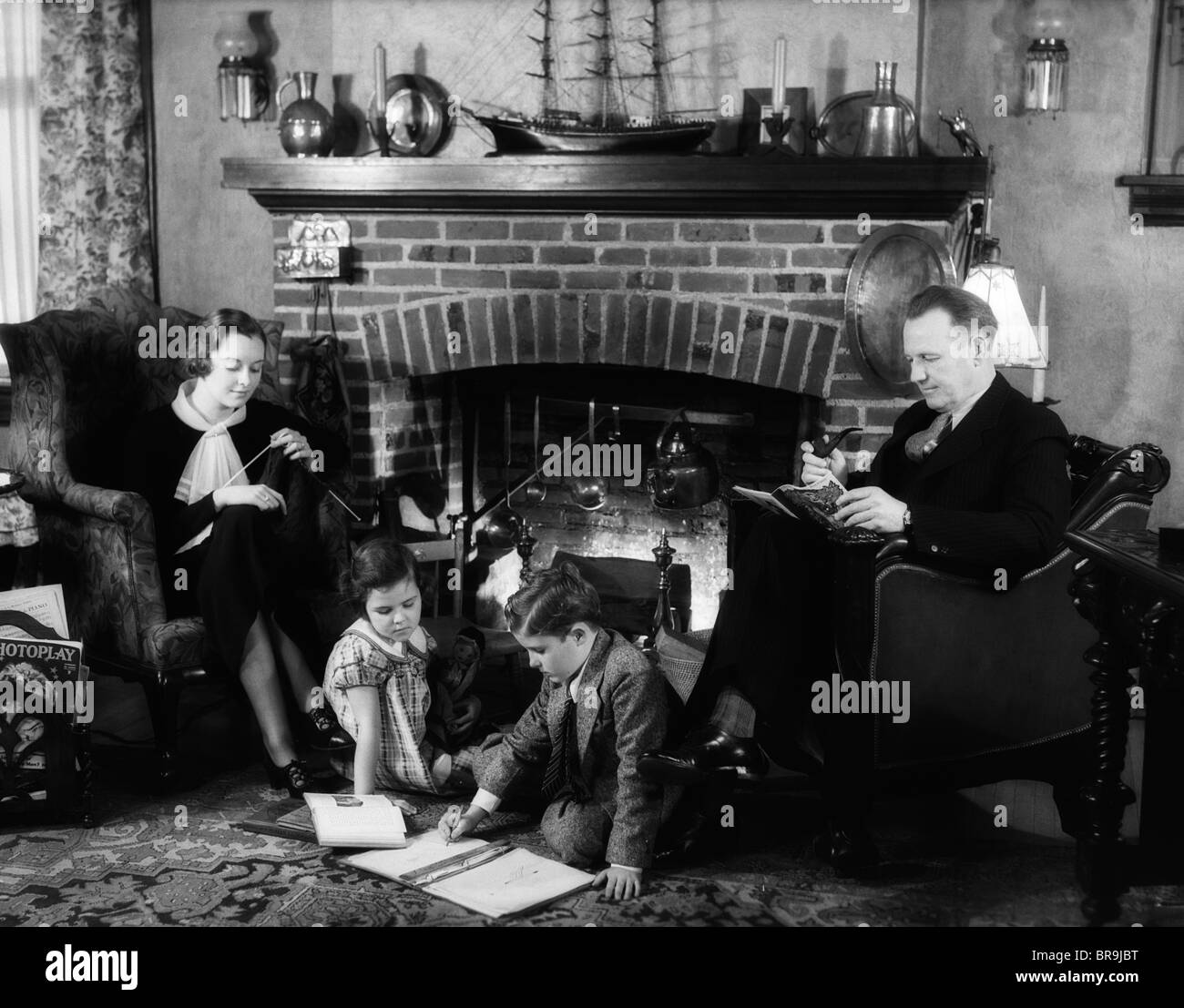 1930s FAMILY OF FOUR SITTING IN FRONT FIREPLACE MOTHER KNITTING FATHER READING SMOKING PIPE KIDS DOING HOMEWORK Stock Photo