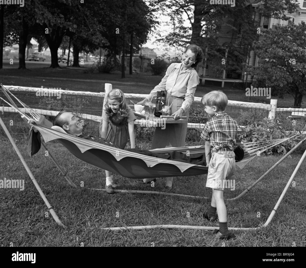 1950s MOM & KIDS SERVING DAD IN HAMMOCK Stock Photo