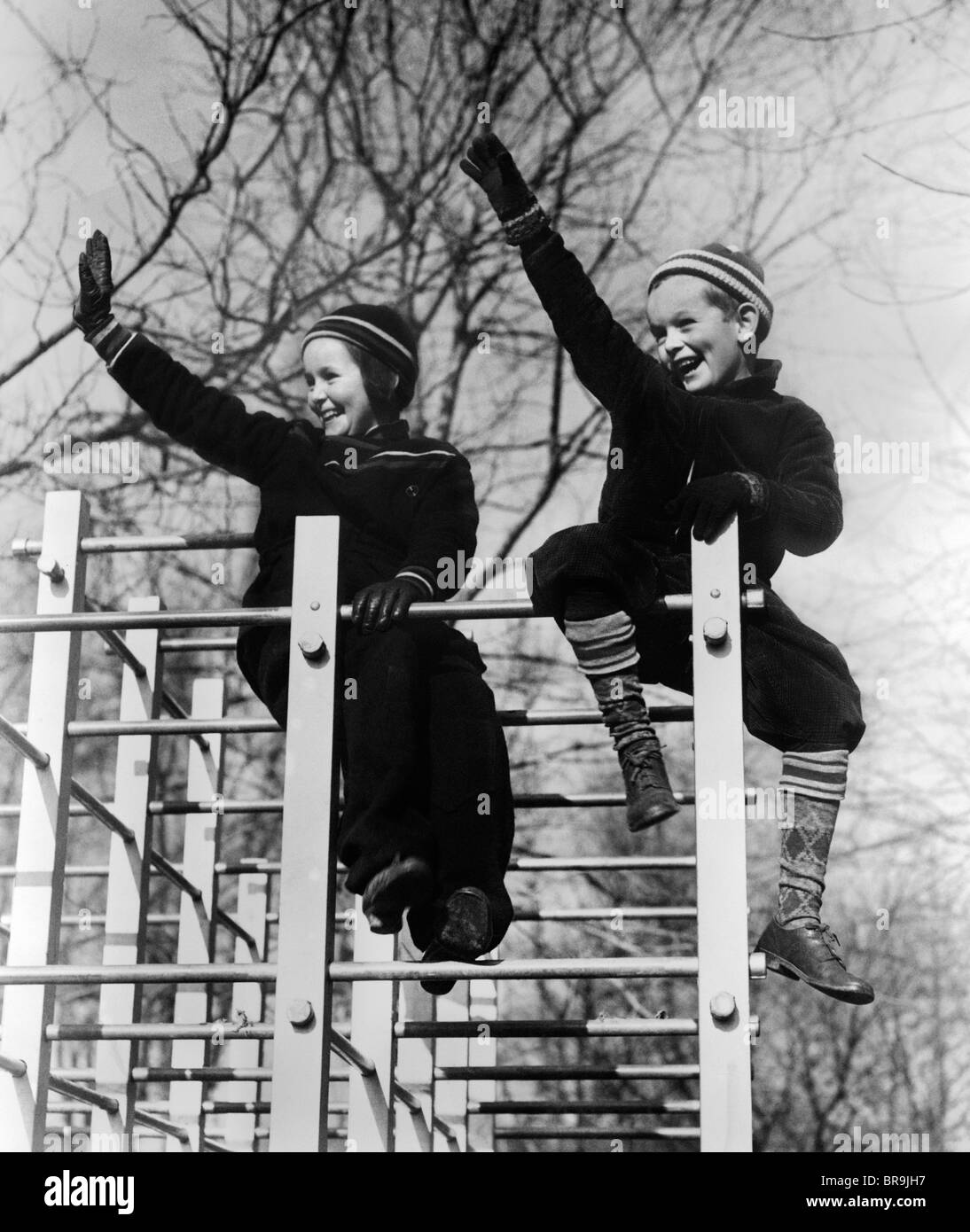 1930s TWO CHILDREN WAVING WHILE SITTING ON PLAYGROUND EQUIPMENT Stock Photo