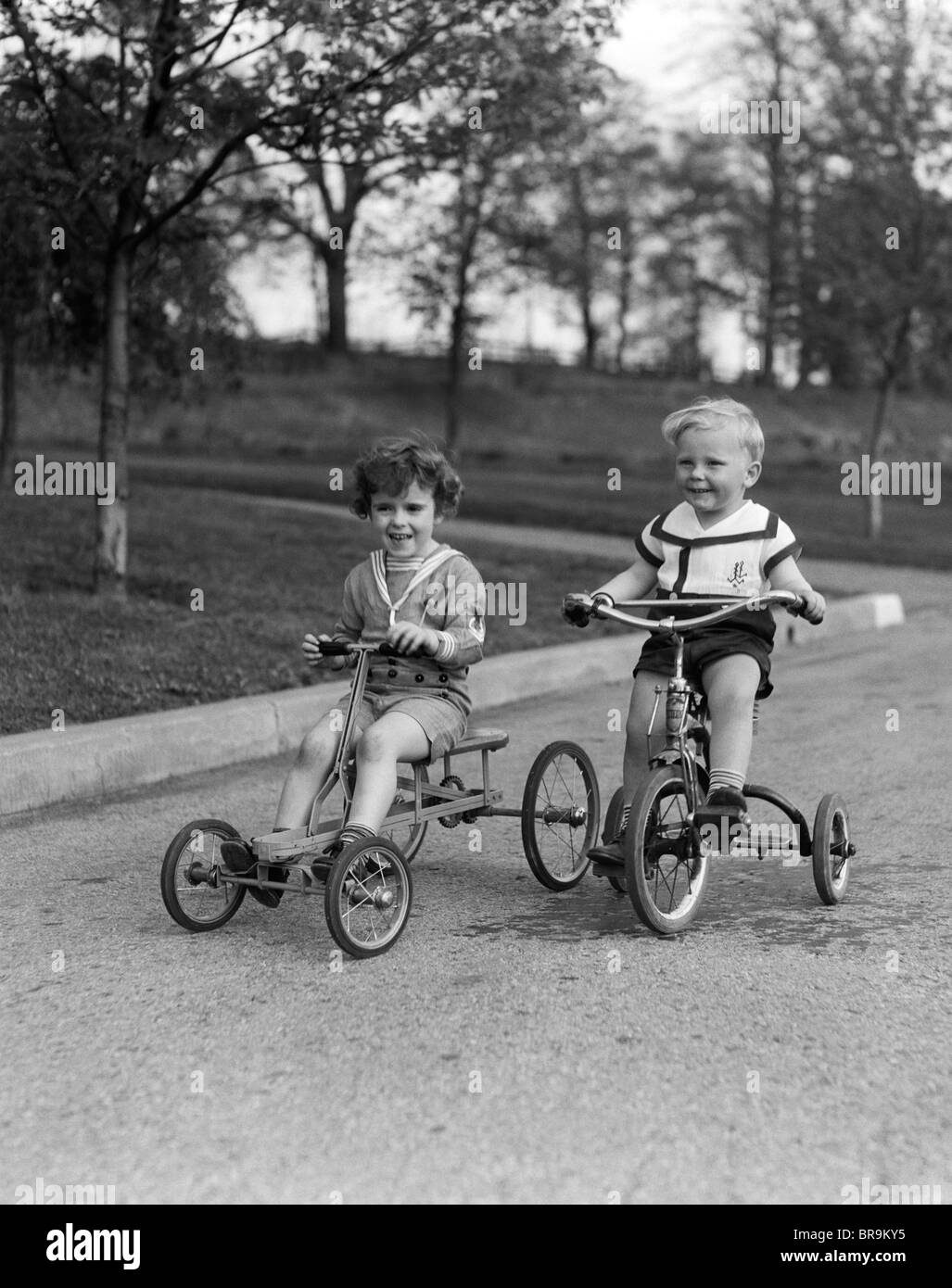 1930s TWO SMILING BOYS RIDING TRICYCLES Stock Photo