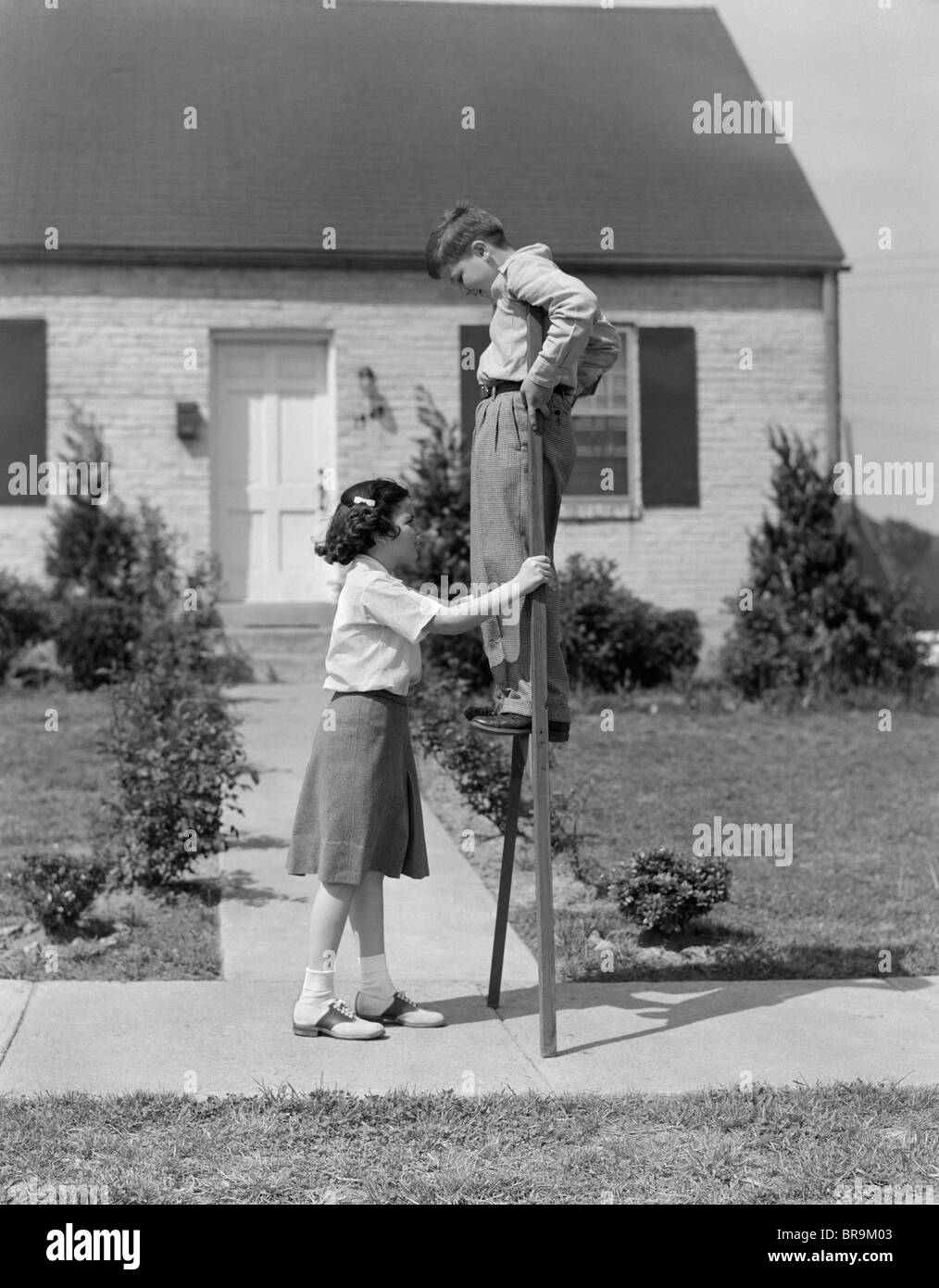 1950s CHILDREN BOY AND GIRL PLAYING WITH STILTS STANDING WALKING ON SIDEWALK Stock Photo