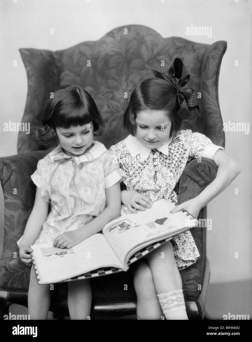 1920s 1930s TWO GIRLS SITTING SIDE BY SIDE IN CHAIR READING BOOK Stock Photo