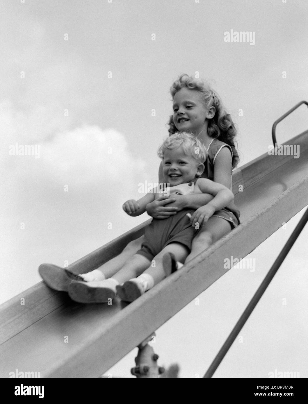1950s 1960s TWO SMILING CHILDREN GOING DOWN PLAYGROUND SLIDE Stock Photo