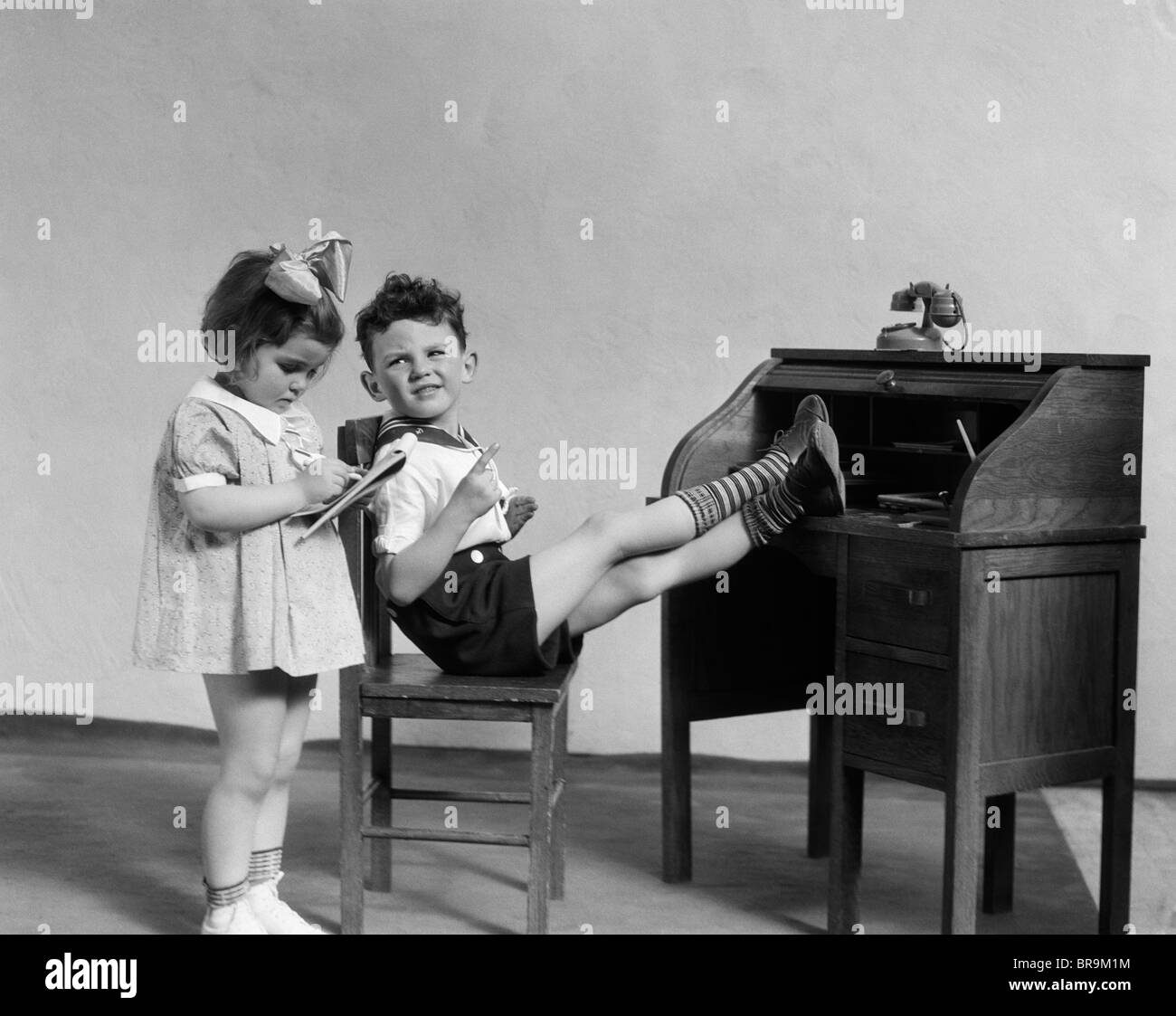 1930s TWO CHILDREN BOY AND GIRL PLAYING OFFICE BOSS FEET ON DESK SECRETARY TAKING DICTATION Stock Photo