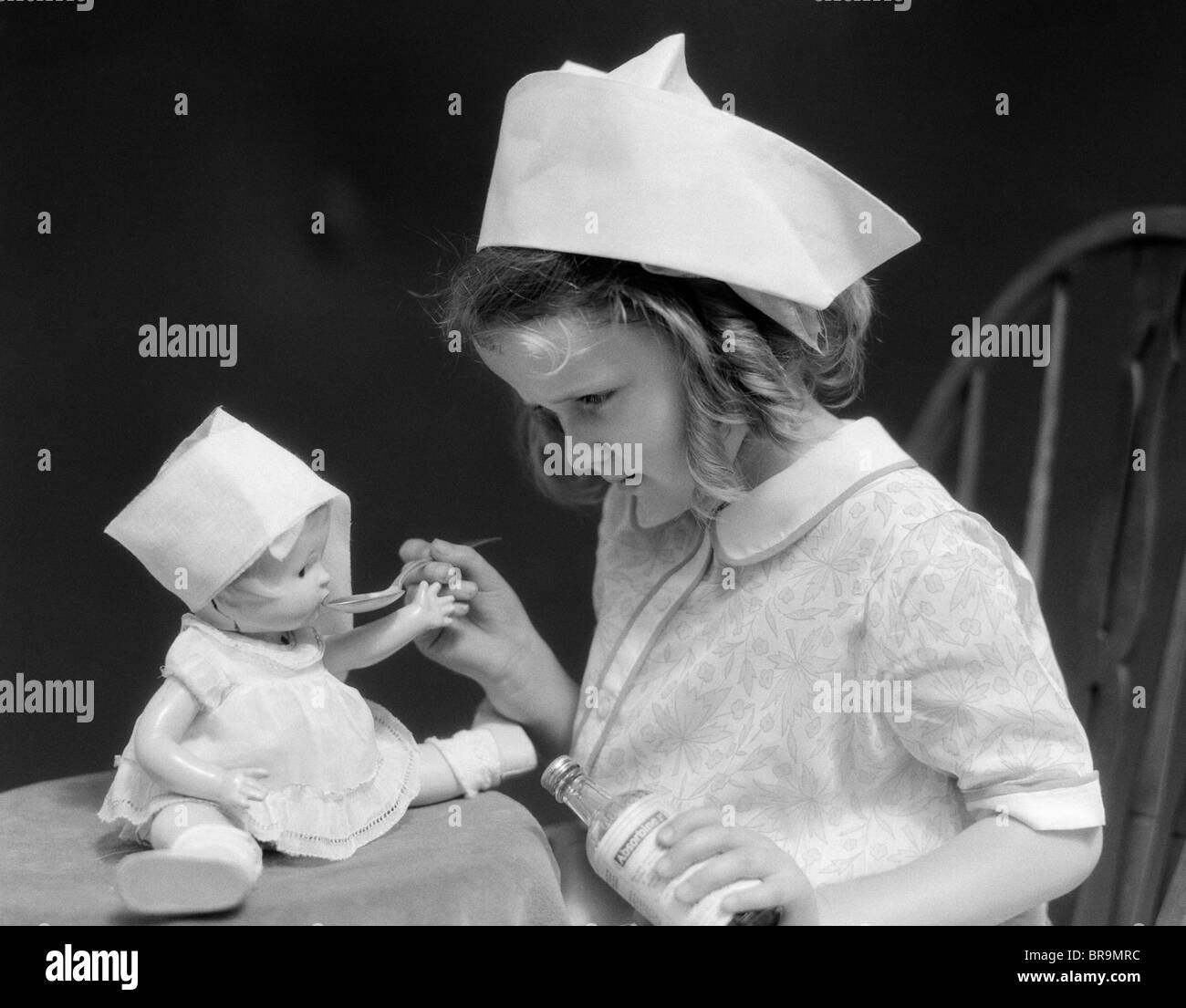 1930s LITTLE GIRL PLAYING NURSE WITH DOLL SPOONFUL OF MEDICINE Stock Photo