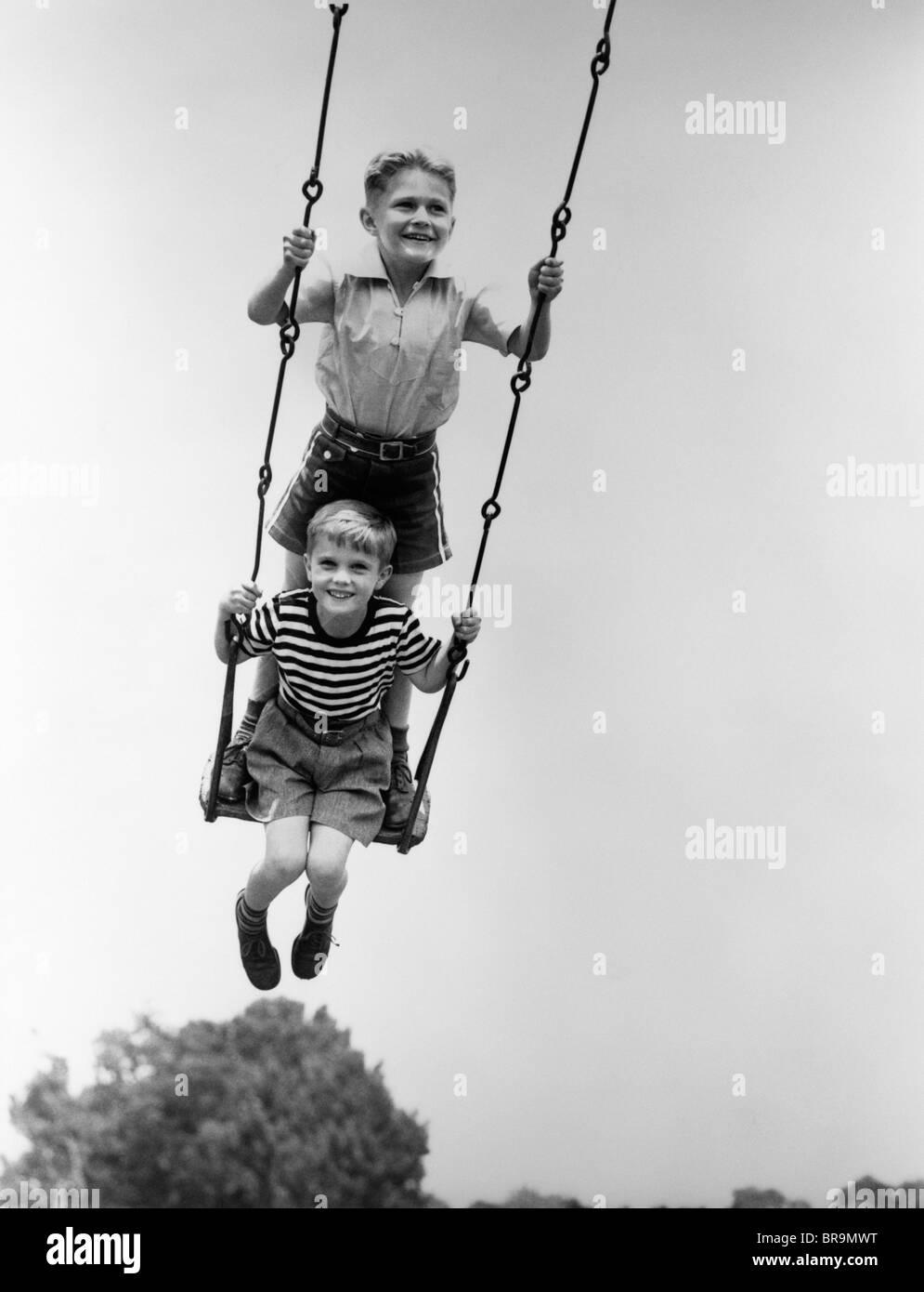 1930s TWO SMILING BOYS SITTING STANDING ON PLAYGROUND SWING Stock Photo