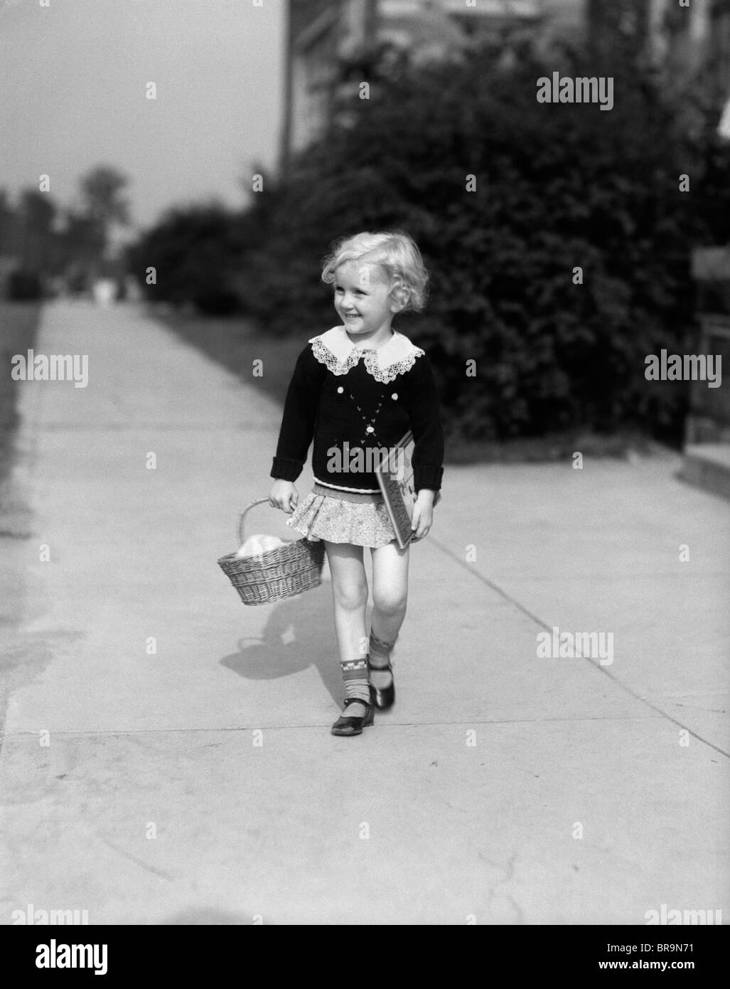 1940 LITTLE BLOND GIRL HOLDING BASKET BOOKS WALKING SIDEWALK Stock Photo