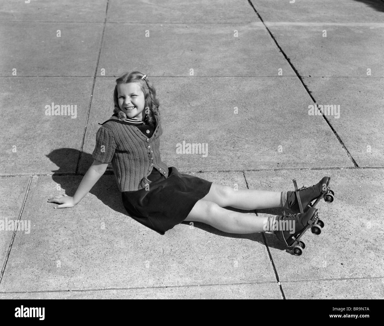 1940s LITTLE GIRL SITTING ON GROUND WEARING ROLLER-SKATES LOOKING AT CAMERA Stock Photo