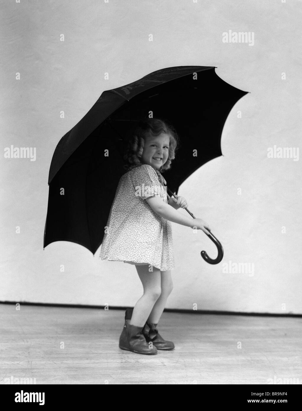 1930s CHILD SMILING LAUGHING LITTLE GIRL WITH CURLY RINGLETS HOLDING LARGE ADULT SIZE UMBRELLA Stock Photo