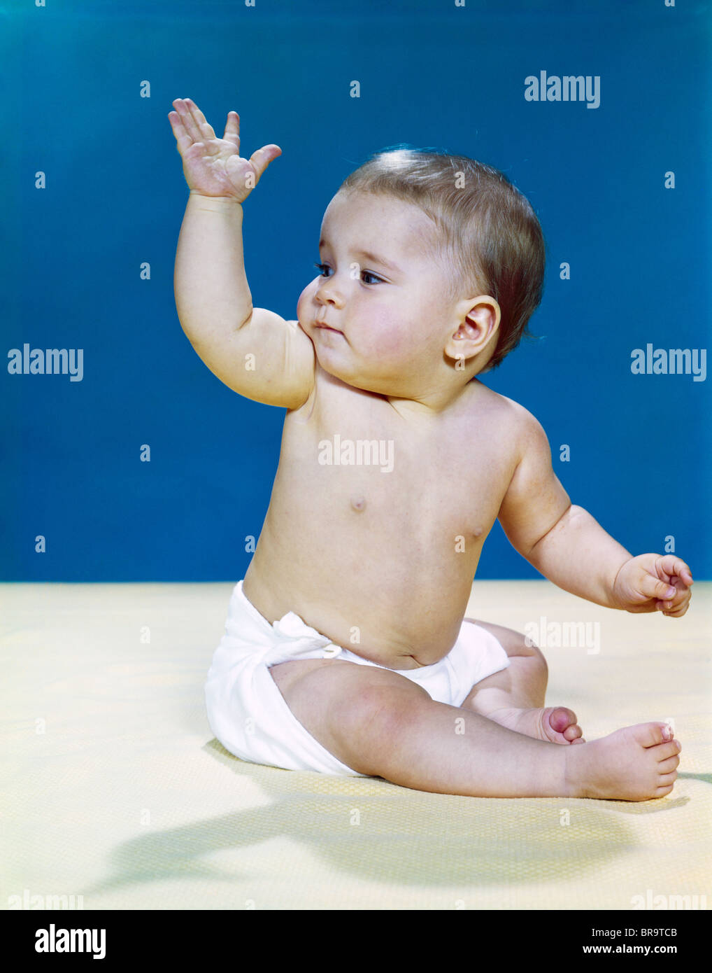 1960s BABY WEARING CLOTH DIAPER LOOKING OVER SHOULDER WAVING WITH RAISED ARM GESTURE Stock Photo
