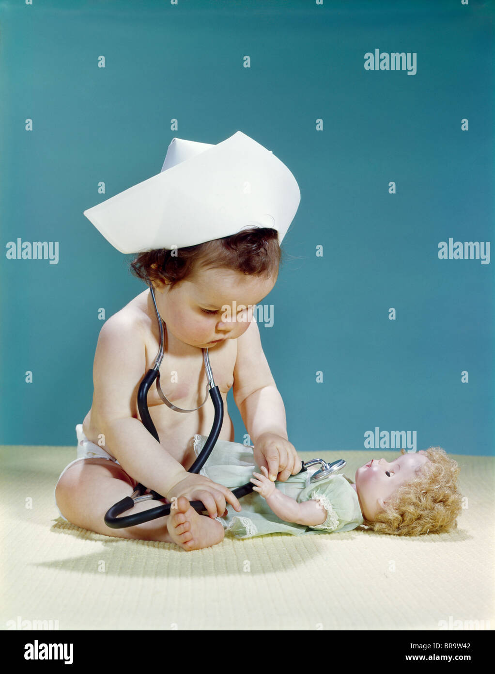 1960s BABY WEARING NURSE NURSE'S CAP STETHOSCOPE LISTENING TO DOLL HEARTBEAT CHEST Stock Photo