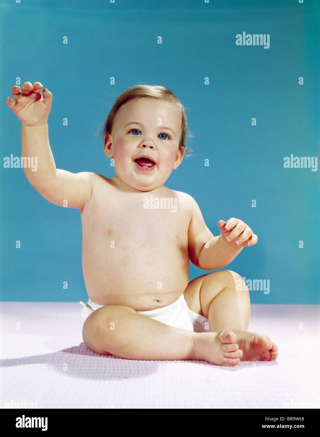 1960s HAPPY SMILING BABY WEARING DIAPER SITTING AND WAVING RAISED ARM LOOKING AT CAMERA Stock