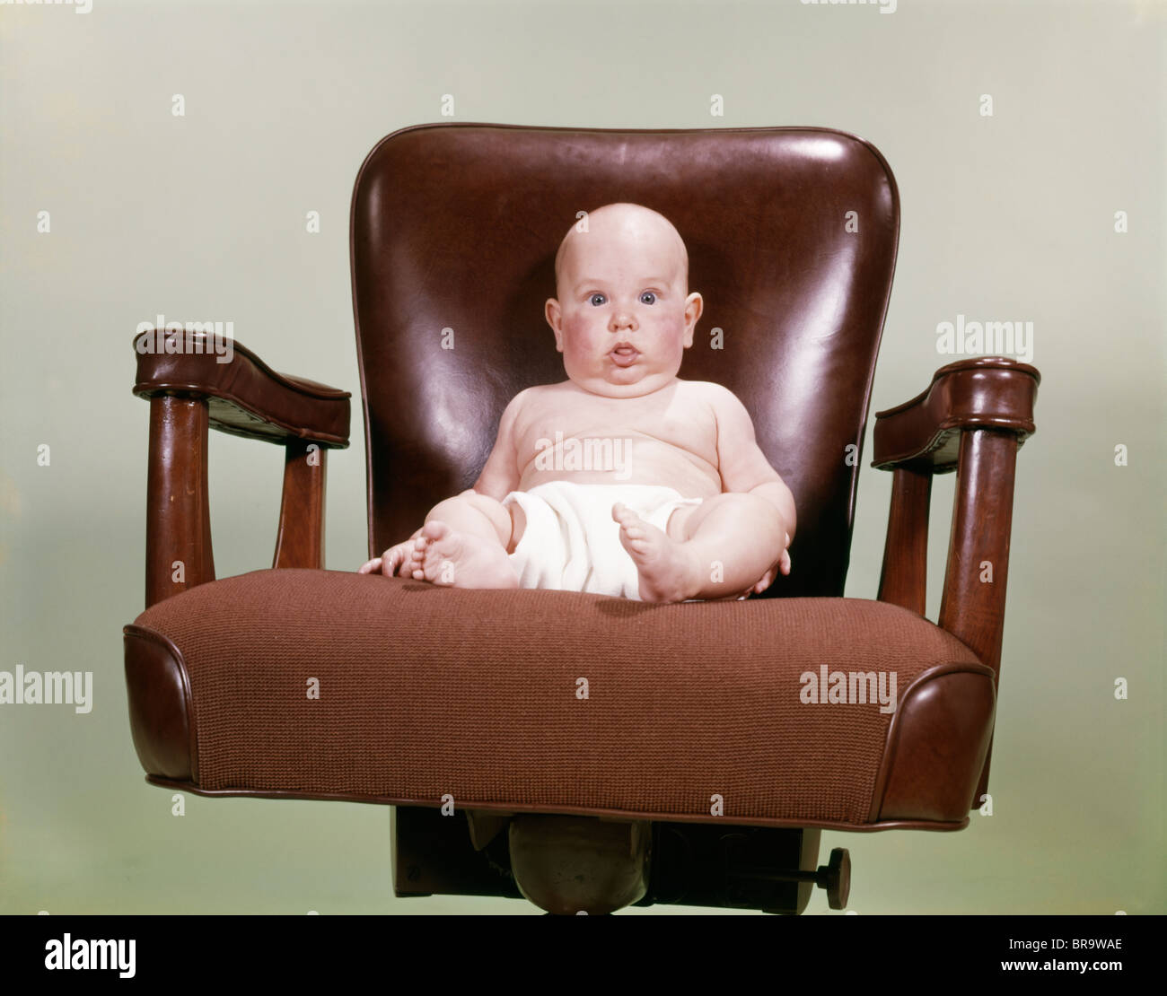 1960s CHUBBY BALD BABY WEARING CLOTH DIAPER SITTING IN EXECUTIVE OFFICE BUSINESS CHAIR Stock Photo