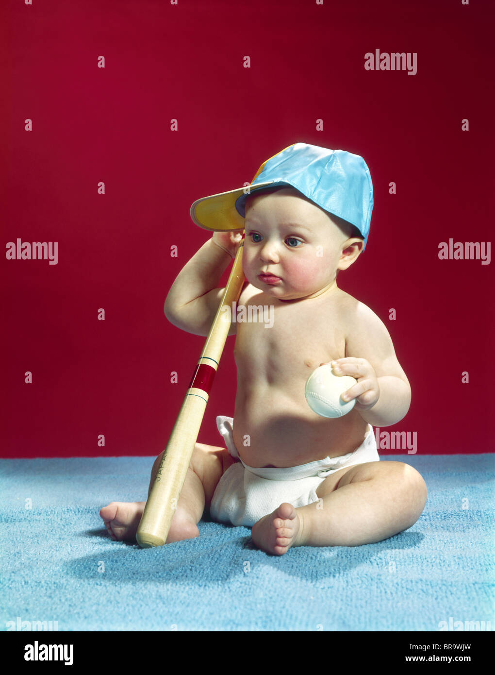 1960s BABY WEARING BLUE BASEBALL CAP HOLDING BALL AND BAT Stock Photo