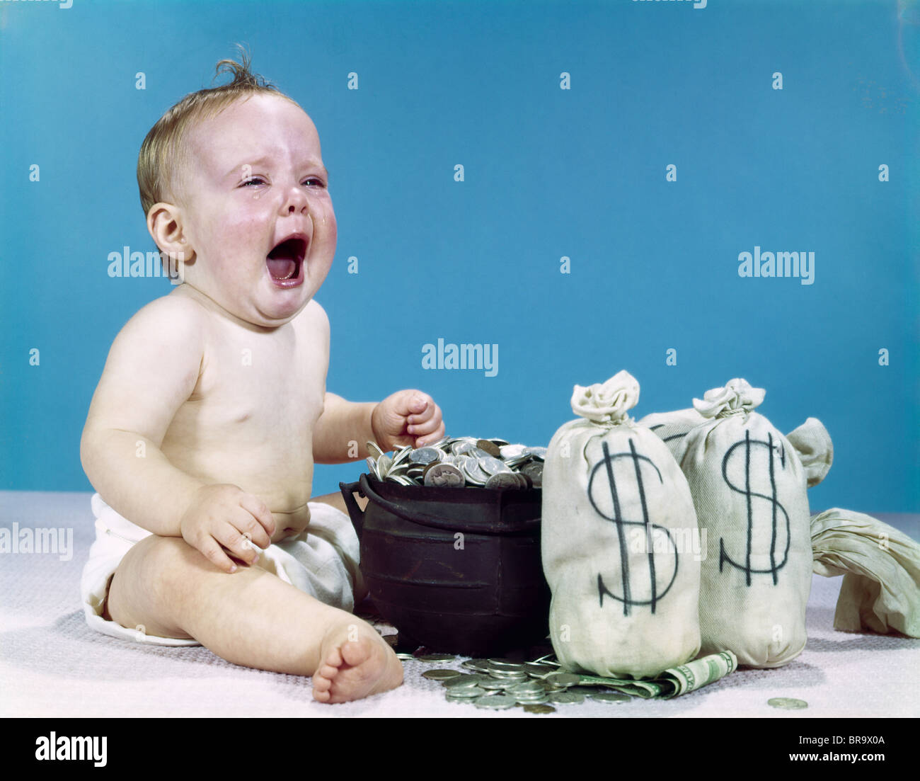 1960s CRYING SHOUTING BABY WITH MONEY BAGS AND POT OF COINS WIN LOTTERY FORTUNE WEALTH TEARS OF JOY Stock Photo