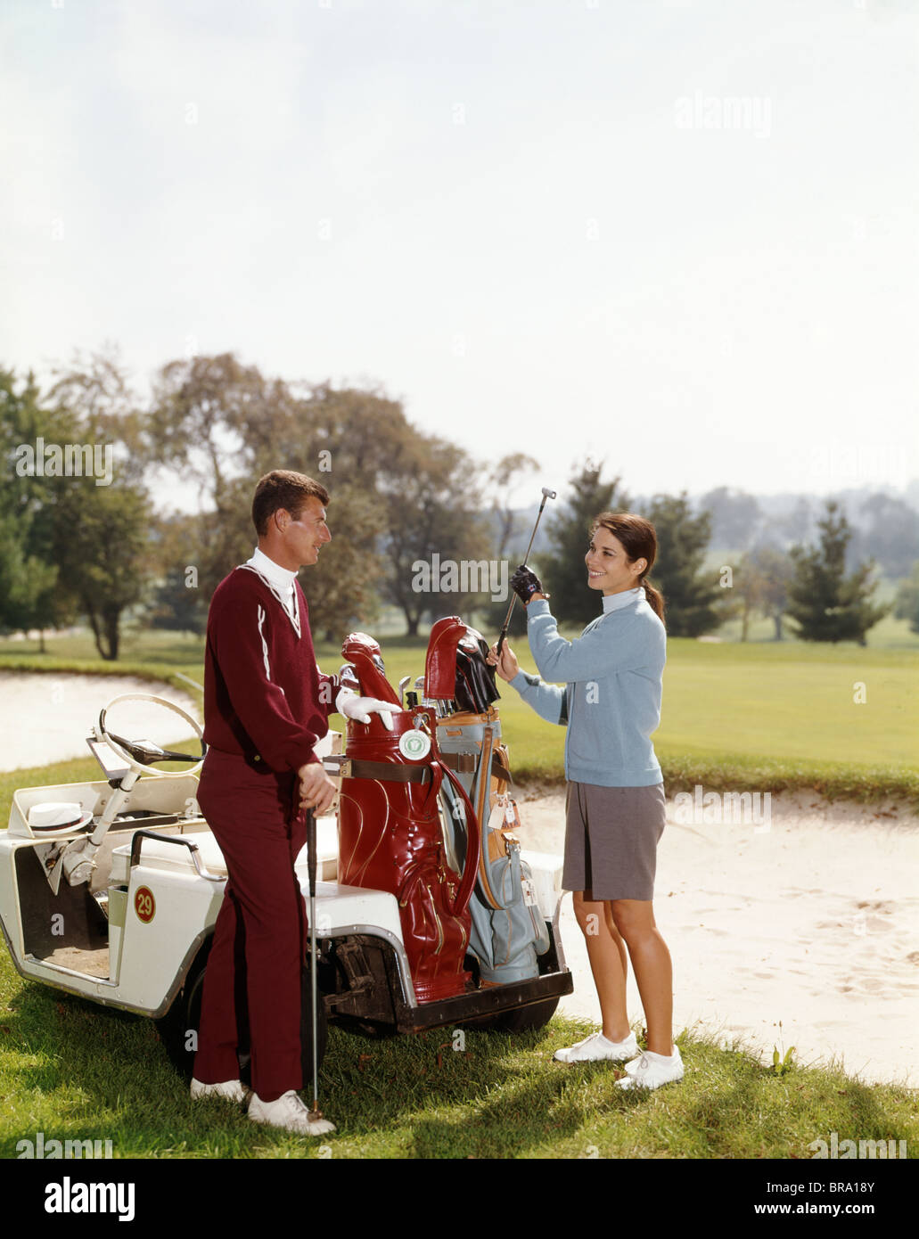 1960s SMILING COUPLE MAN WOMAN SELECTING GOLF CLUBS FROM GOLF BAG ON CART BY SAND TRAP Stock Photo