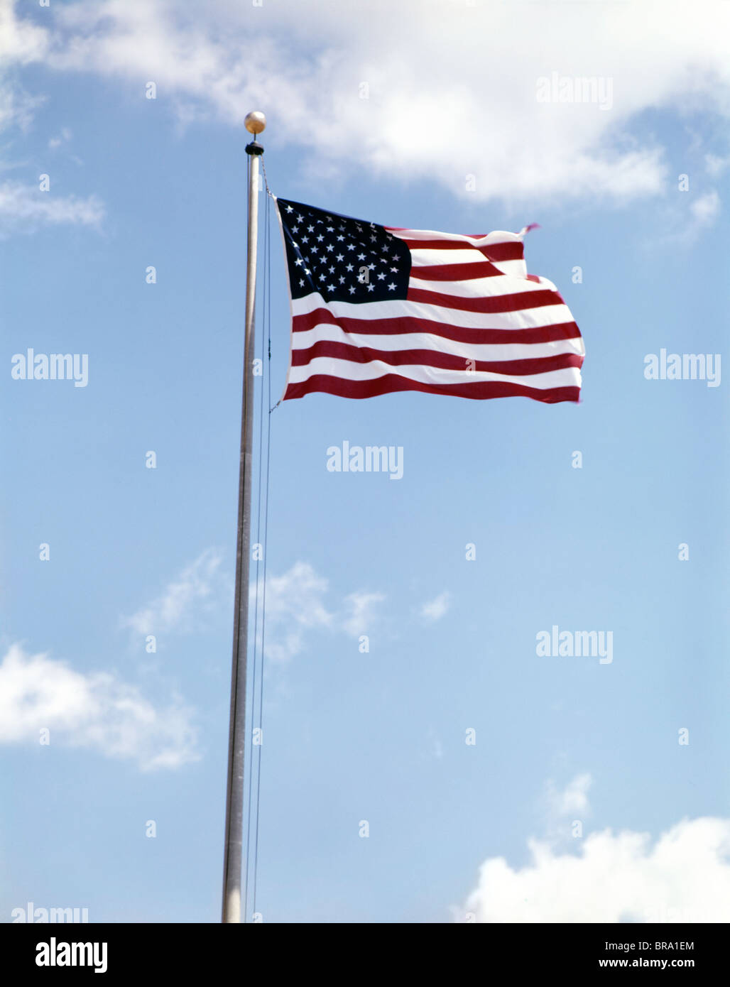 1960s AMERICAN FLAG ON POLE FLYING AGAINST BLUE SKY WITH CLOUDS Stock Photo