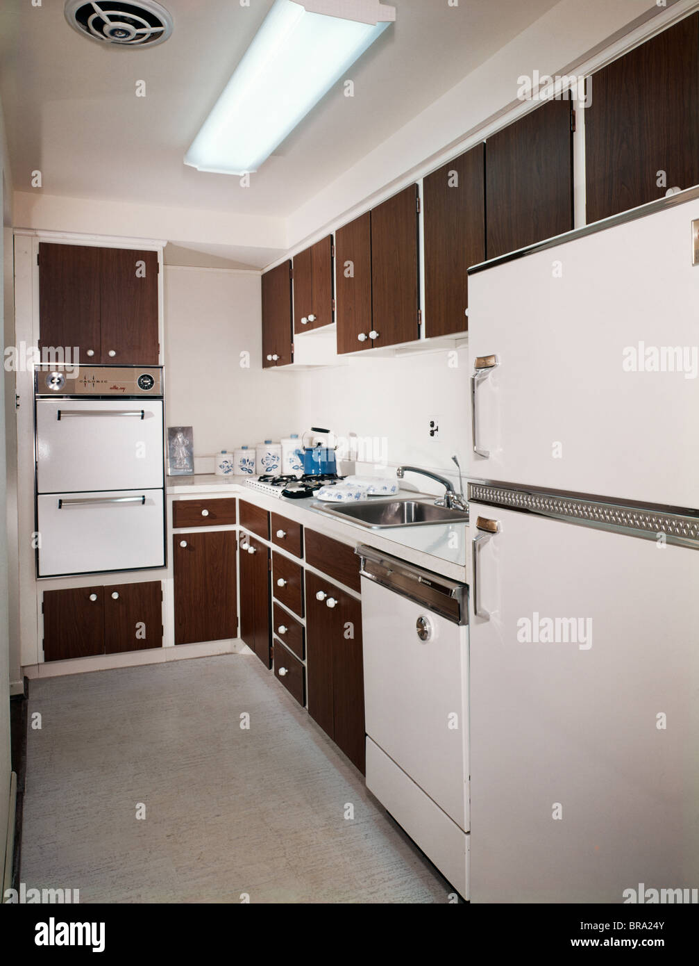 1970s NARROW GALLEY STYLE KITCHEN WITH DARK WOODEN CABINETS AND WHITE APPLIANCES Stock Photo