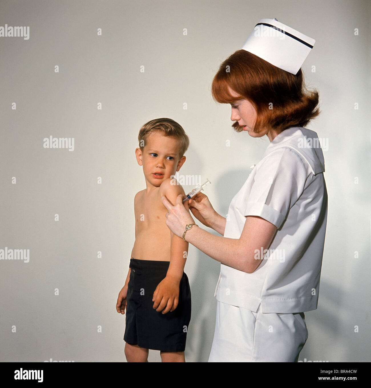 1960s WOMAN NURSE GIVE INJECTION NEEDLE TO LITTLE BOY ARM VACCINE VACCINATION PREVENTION IMMUNIZE INOCULATE MEDICINE Stock Photo