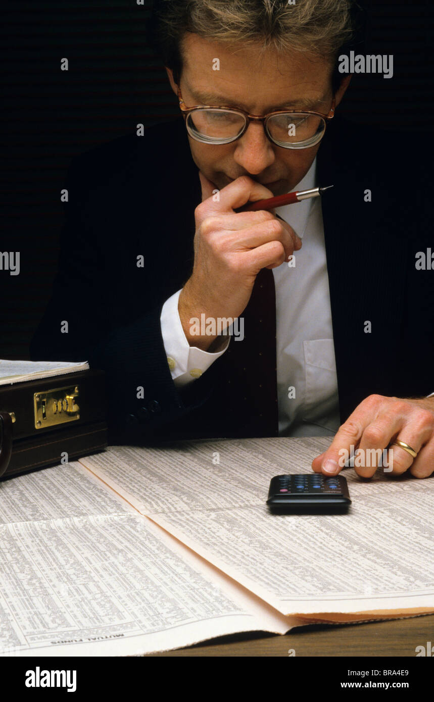 1980 1980s MAN STUDY FINANCIAL SECTION OF NEWSPAPER USING CALCULATOR Stock Photo