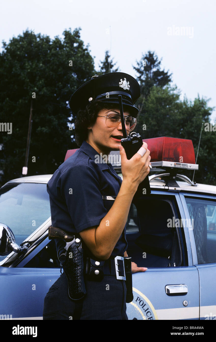 1970s POLICE WOMAN TALKS ON WALKIE TALKIE IN FROMT OF POLICE CAR Stock Photo