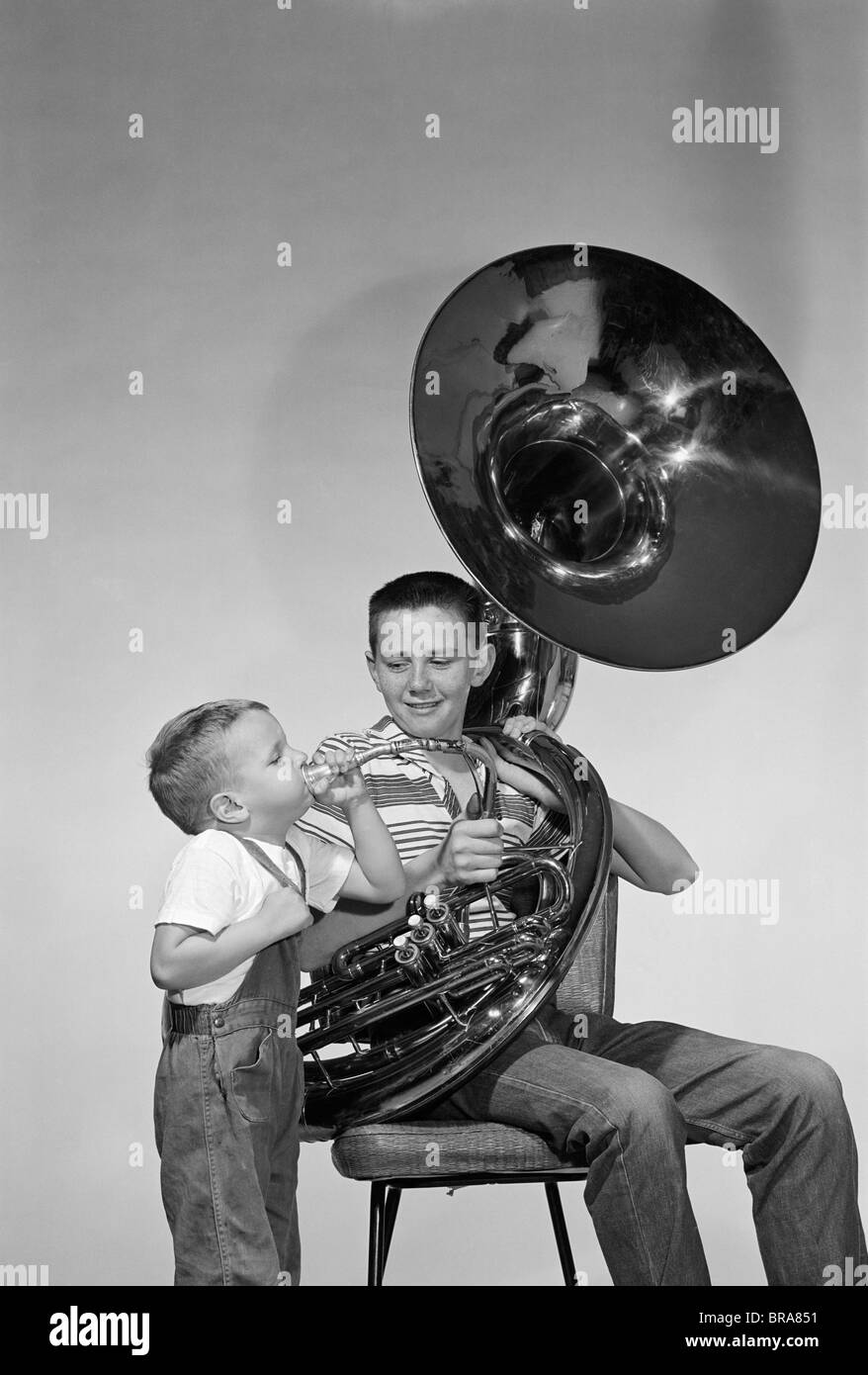 1950s SMILING TEEN BOY WITH BRASS TUBA WEAR STRIPE TEE SHIRT JEANS YOUNGER BOY STANDING BY BLOWING INTO MOUTHPIECE Stock Photo