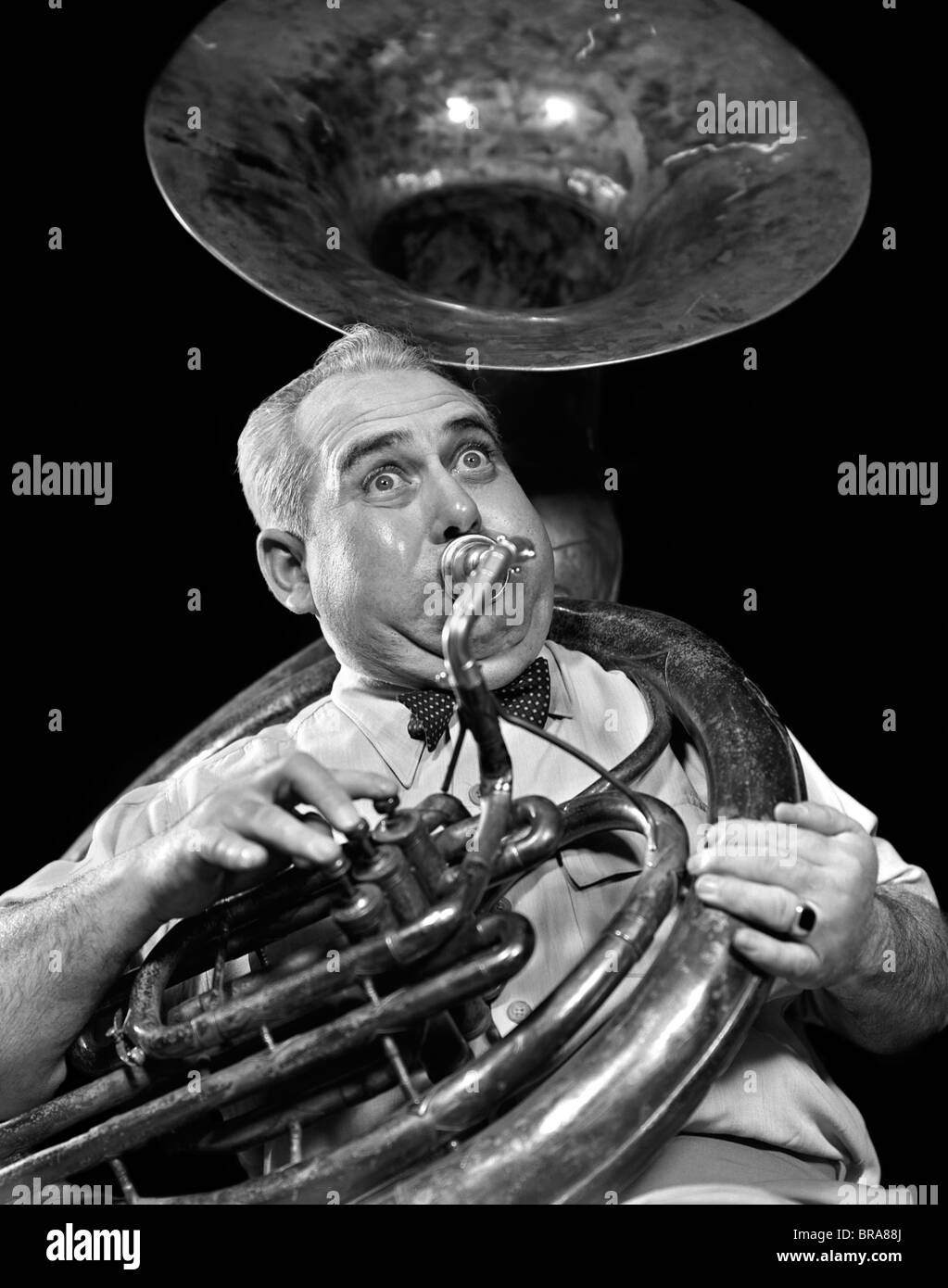 1940s CHUBBY MAN MUSICIAN WITH POLKA DOT BOW TIE AND BULGING EYES PLAYING A SOUSAPHONE Stock Photo