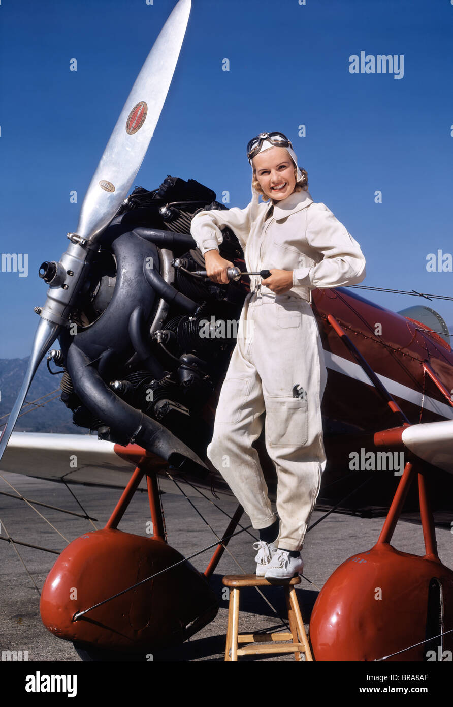 1930s 1940s SMILING WOMAN AIRPLANE PILOT MECHANIC IN COVERALLS STAND WORKING ON PROPELLER ENGINE LOOKING AT CAMERA Stock Photo